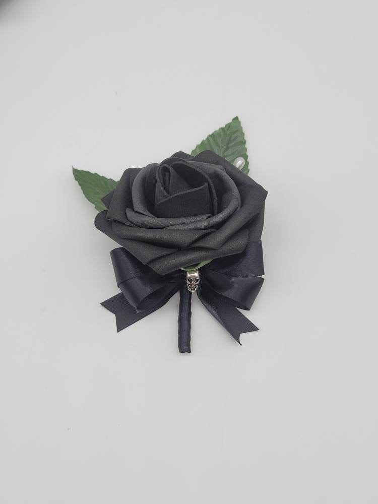 All black boutonniere with black real touch rose and black satin ribbon and finished with a silver skull on stem. also has faux leaves and pearl pin