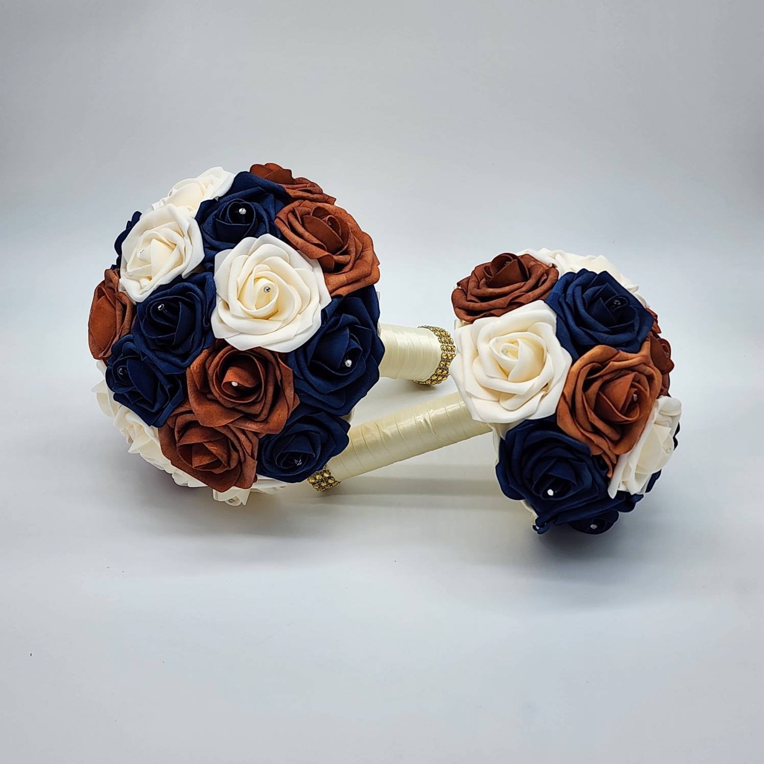 Two bouquets of artificial flowers in shades of ivory, burnt orange, and navy blue. The bouquets are tied with ivory ribbon and have gold accents.