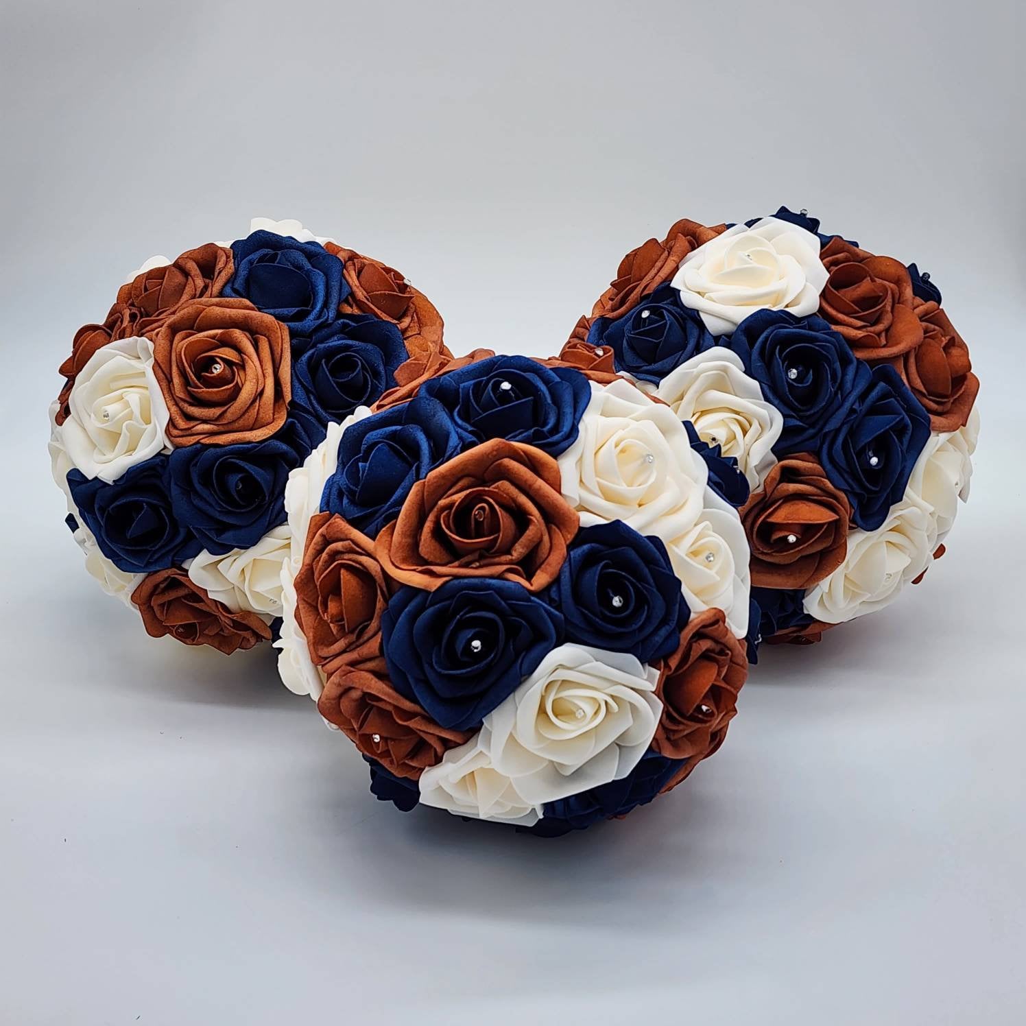 Three round bouquets of artificial flowers. The bouquets are made of ivory, navy blue, and burnt orange-colored roses. The roses are arranged in a circular pattern.