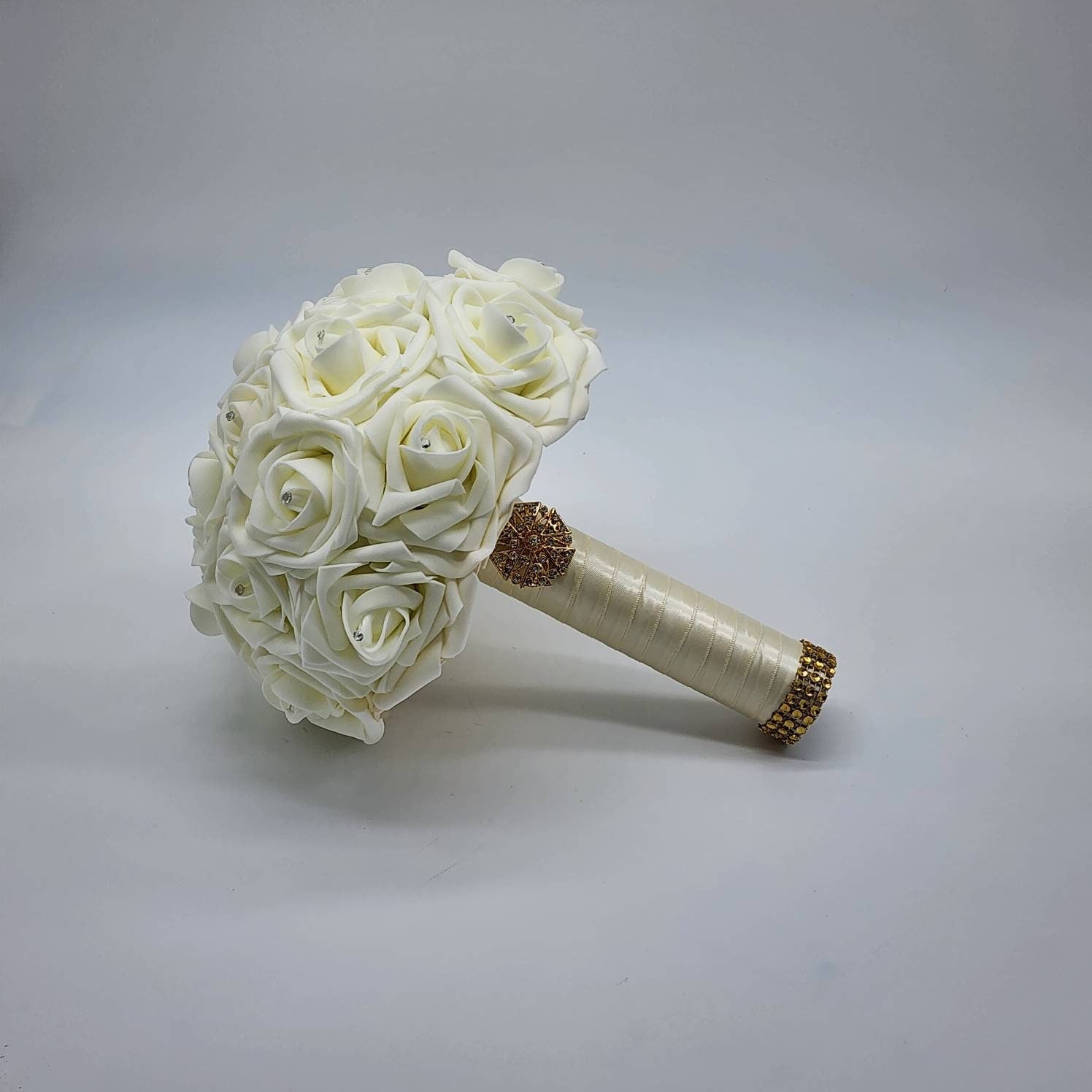 A ivory bridal bouquet made of artificial roses with a gold and rhinestone embellishment on the handle.