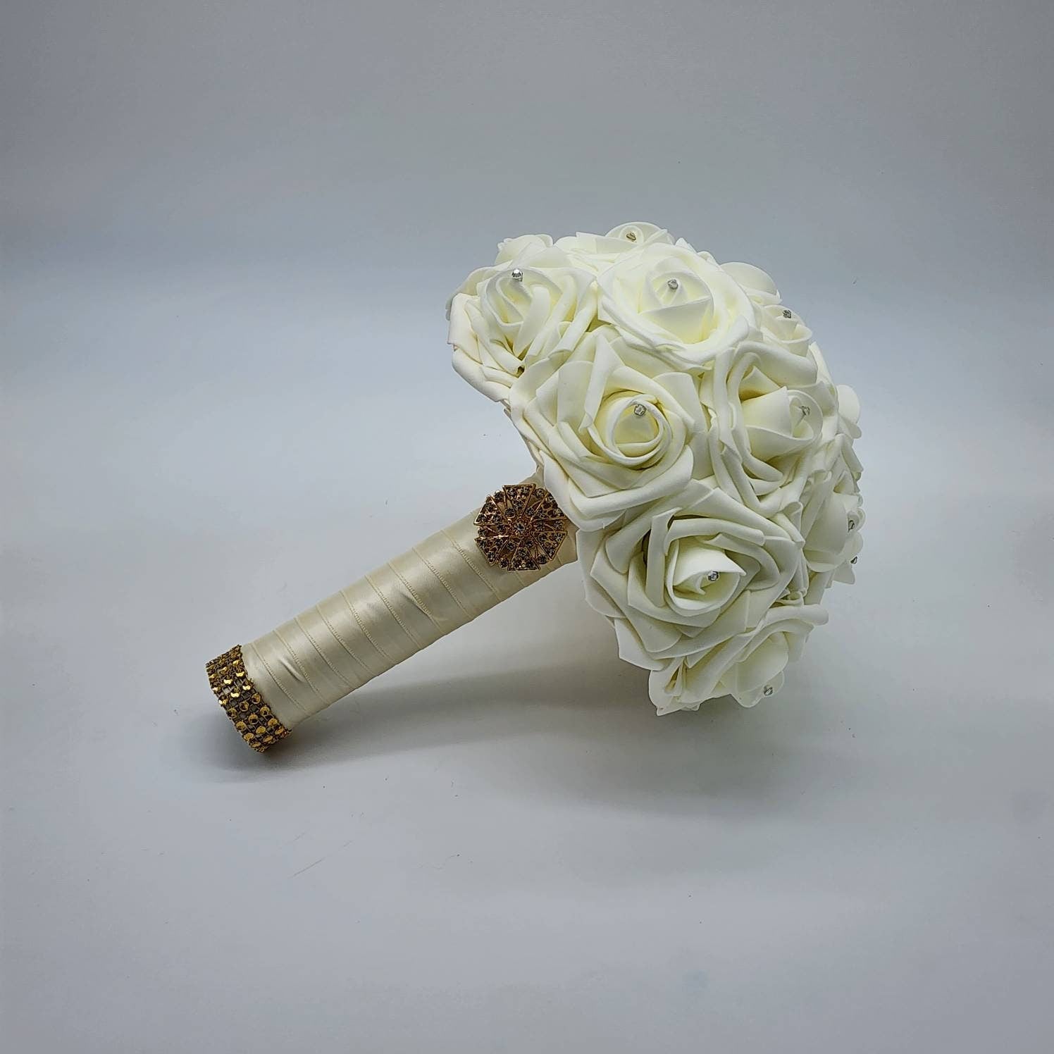 A ivory bridal bouquet made of artificial roses with rhinestone accents and a gold rhinestone embellishment on the handle.