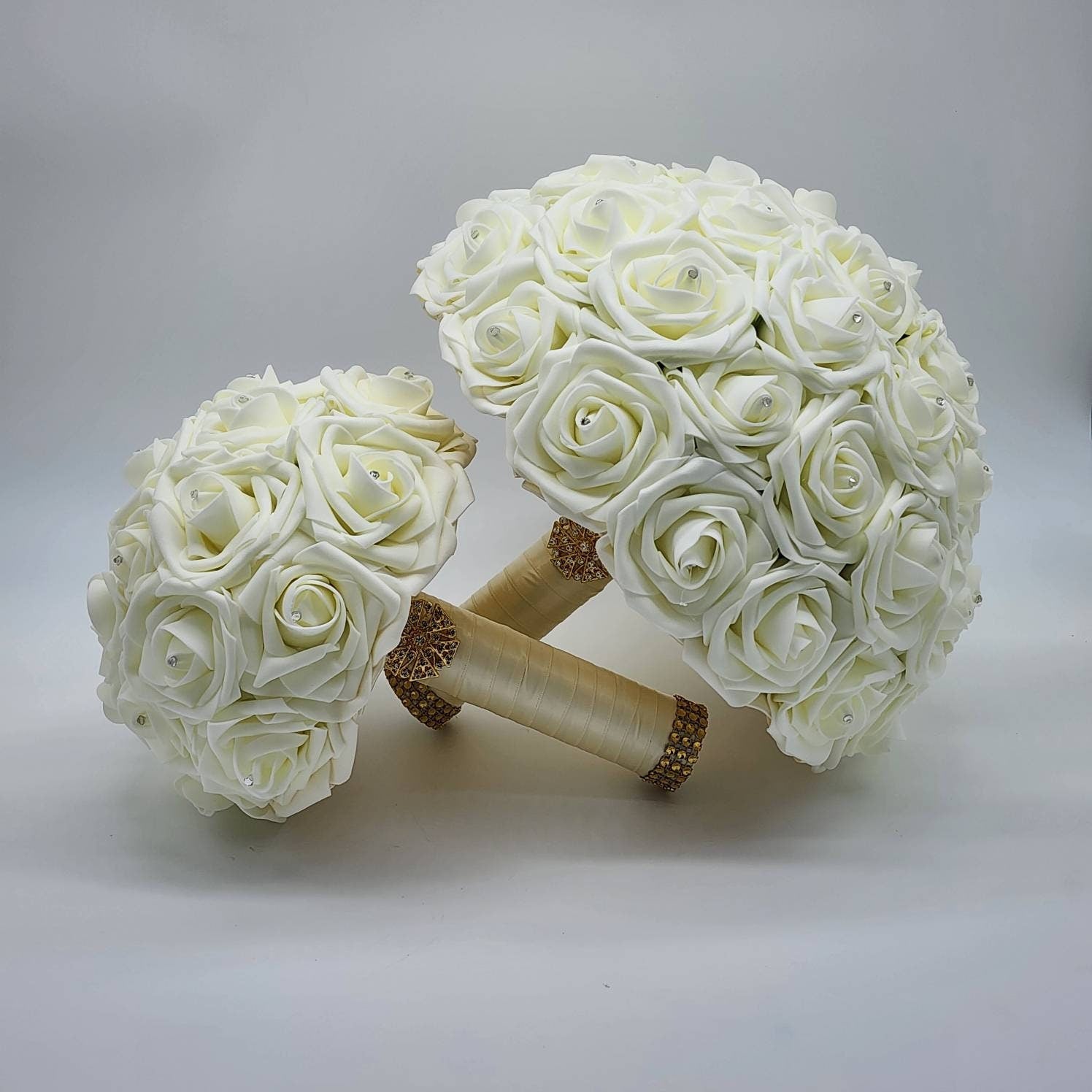 Two ivory faux rose bouquets with gold and rhinestone accents. The larger bouquet is in the background, and the smaller bouquet is in the foreground.
