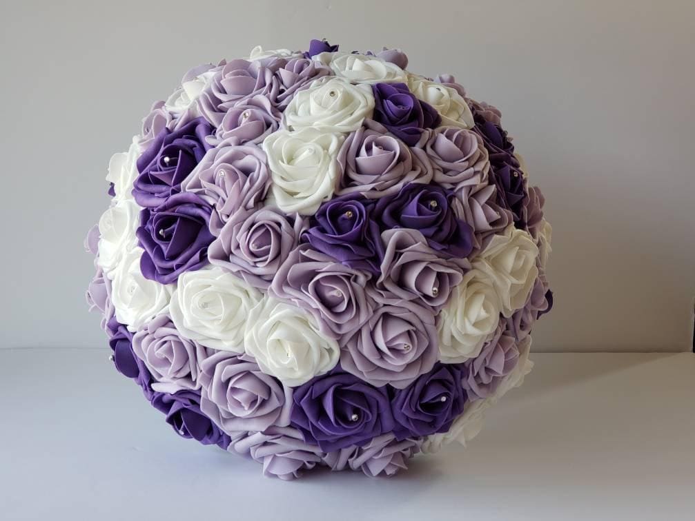 A round bouquet of artificial flowers in shades of purple and white. The bouquet is made of Real Touch roses and is perfect for a wedding or special occasion.