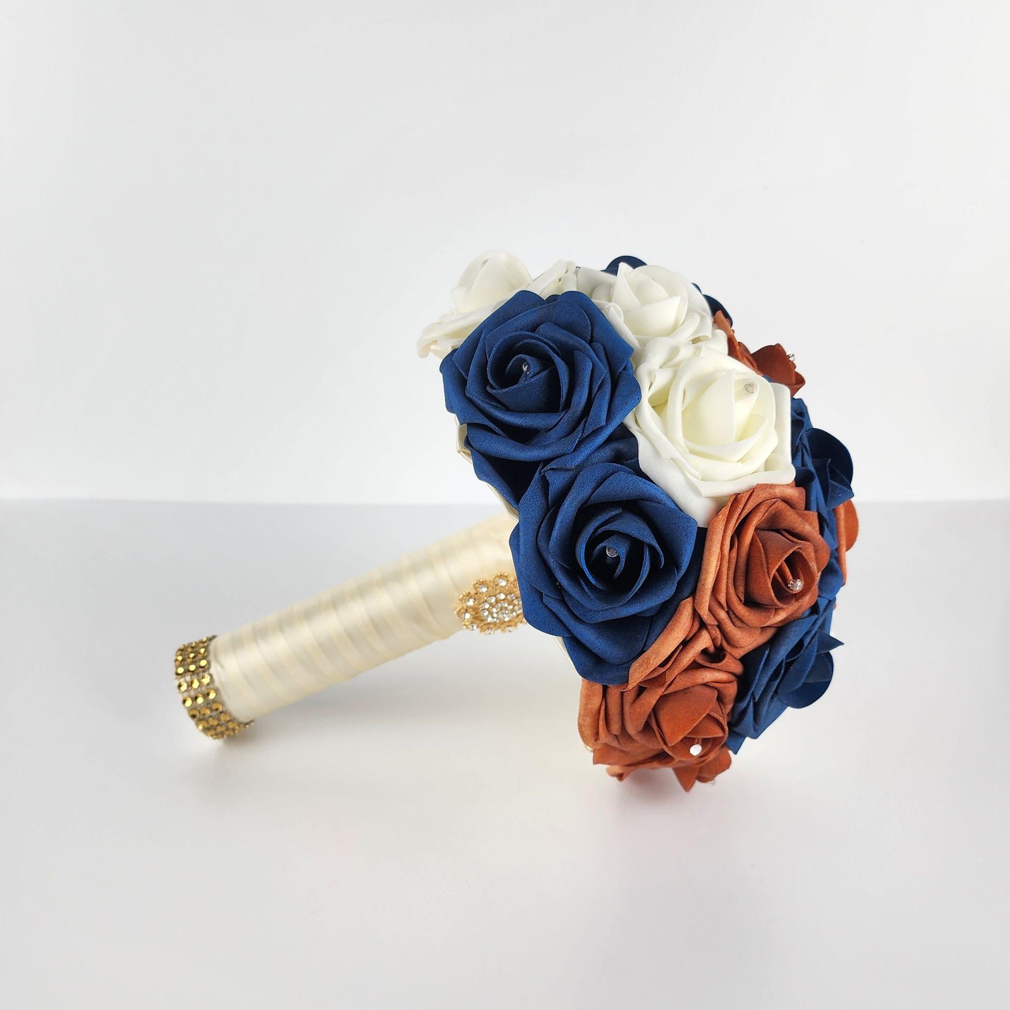 A bridal bouquet with ivory, navy blue, and burnt orange roses. The bouquet has a satin ribbon handle with a gold rhinestone embellishment.