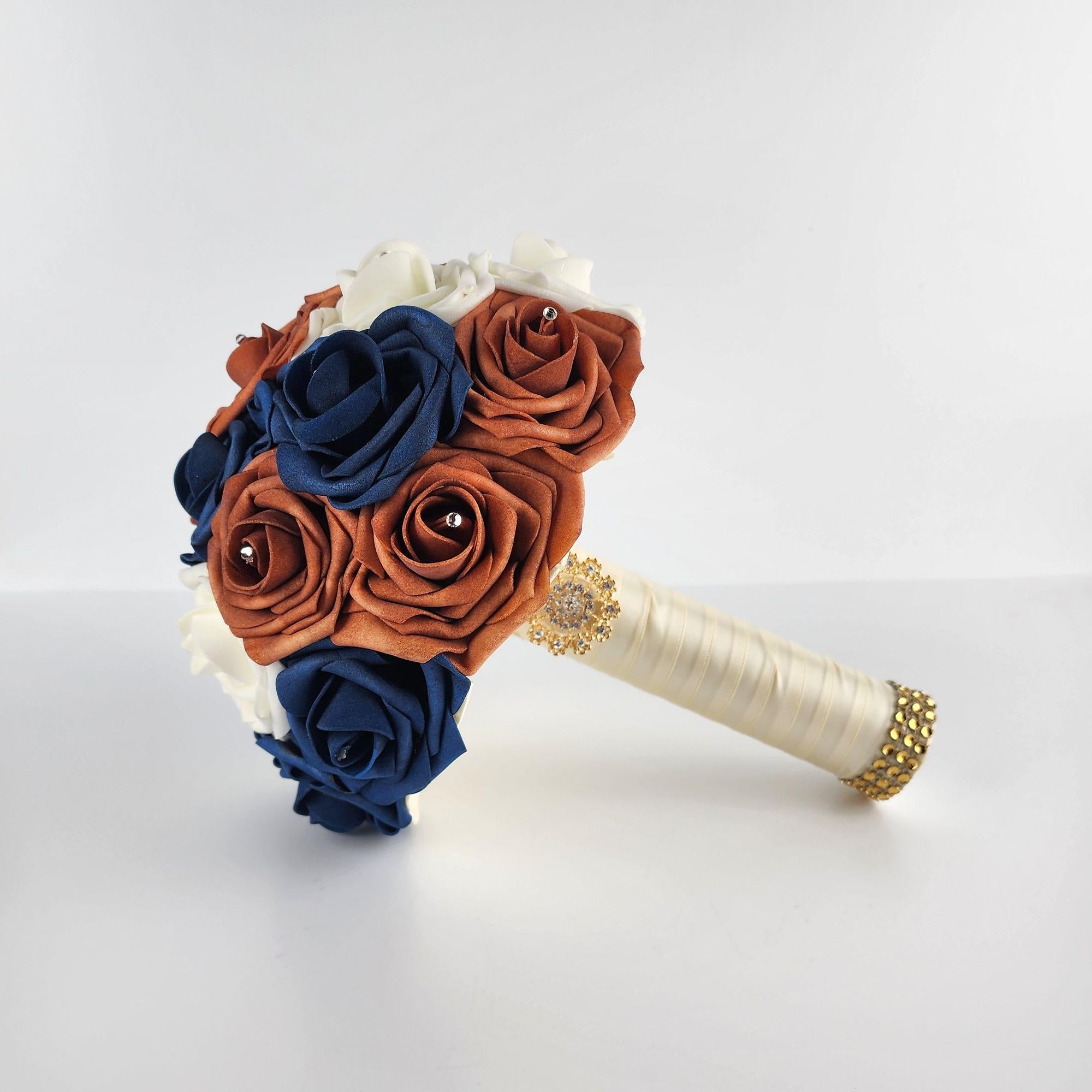 A bridal bouquet with navy blue, burnt orange, and white real touch roses. The bouquet has a white satin ribbon and a gold rhinestone embellishment.