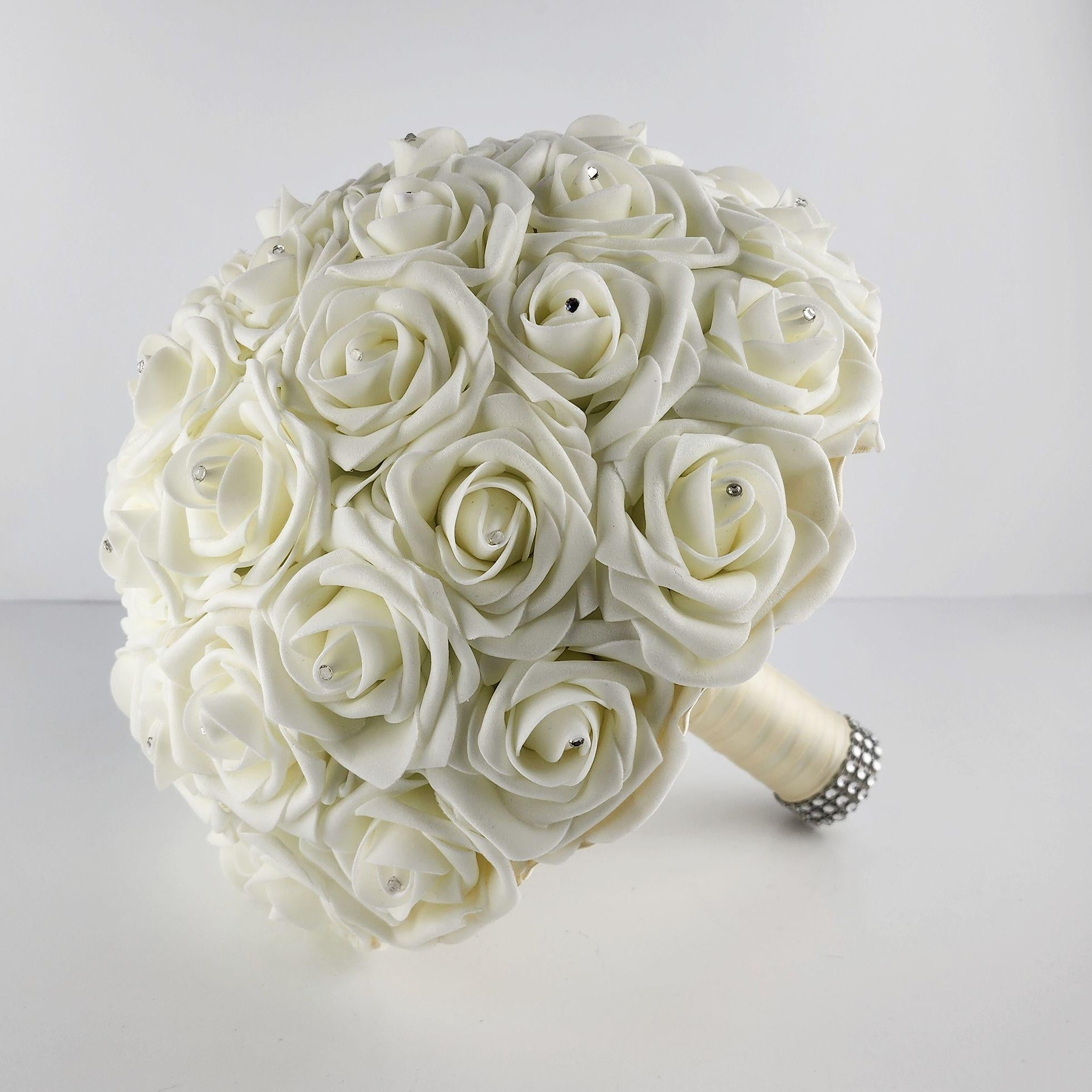 A ivory bridal bouquet made of artificial roses with rhinestones. The bouquet is round and has a satin ribbon handle.