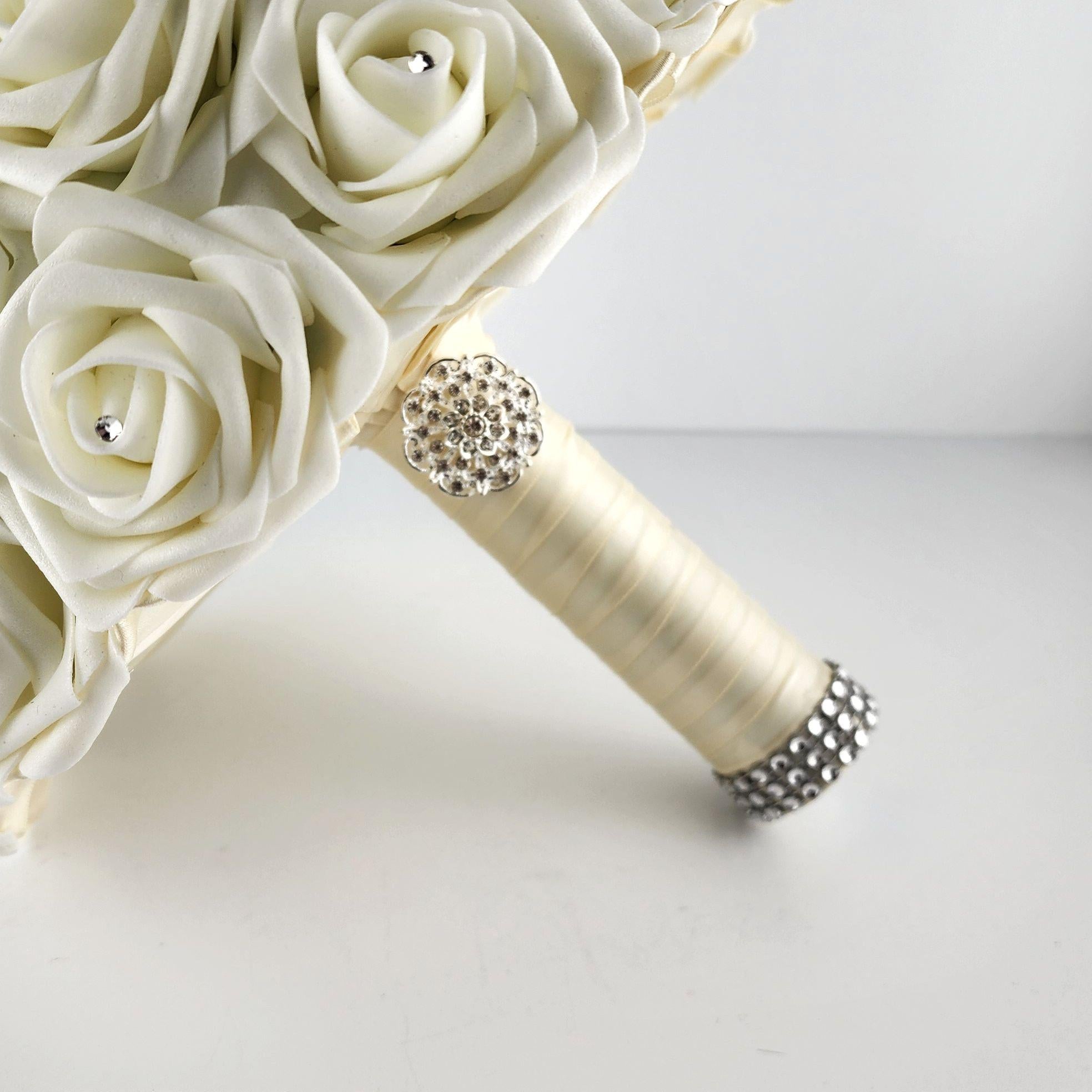A ivory bridal bouquet with a satin ribbon handle and a rhinestone embellishment. The bouquet is made of artificial roses.