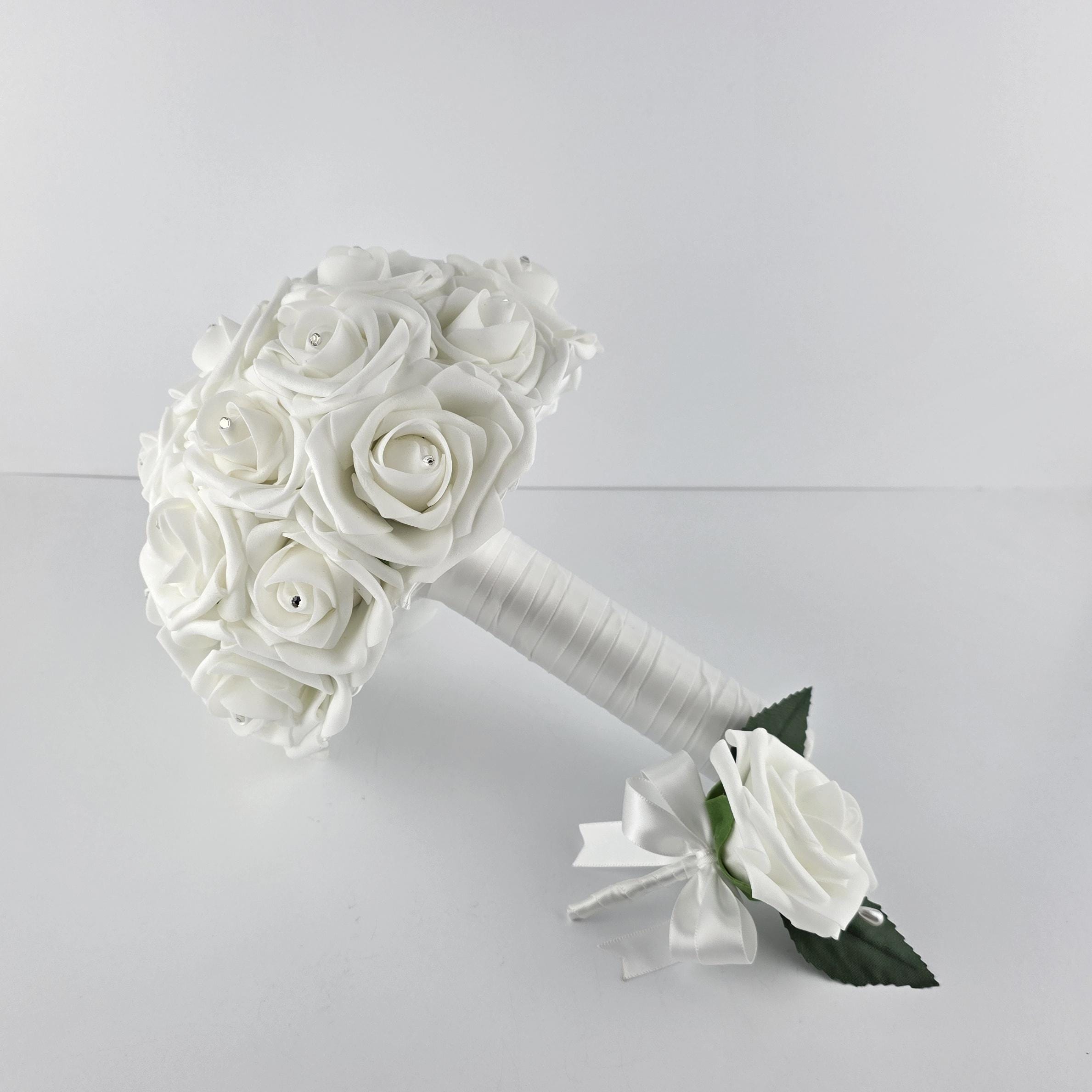 A white bridal bouquet with a matching boutonniere. The bouquet features several white roses with rhinestone accents. The stems are wrapped in white satin ribbon. The boutonniere has a single white rose with a white ribbon.