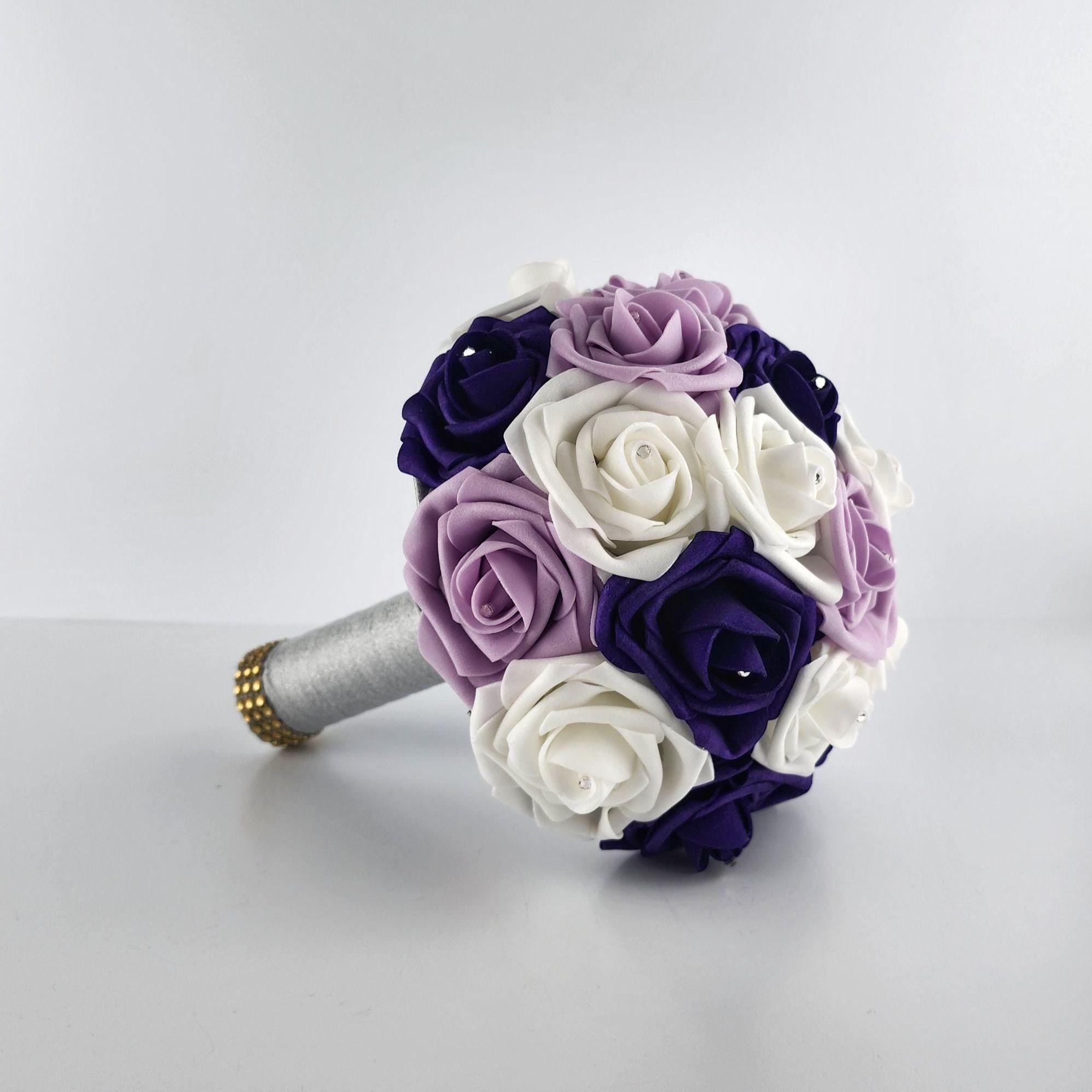 A bouquet of artificial flowers in shades of purple, lavender, and white. The flowers are arranged in a round shape and have a silver stem with gold accents.