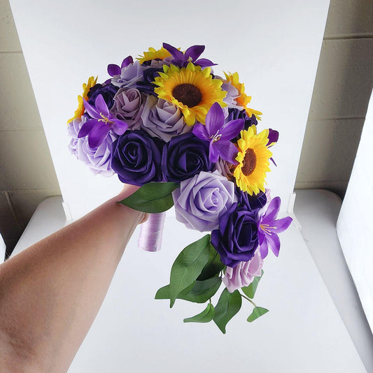 cascading Lilac, lavender, and dark purple real touch roses bridal bouquet with a mix of sunflowers and lilies, and greenery. Handle is made out of lavender silk ribbon.