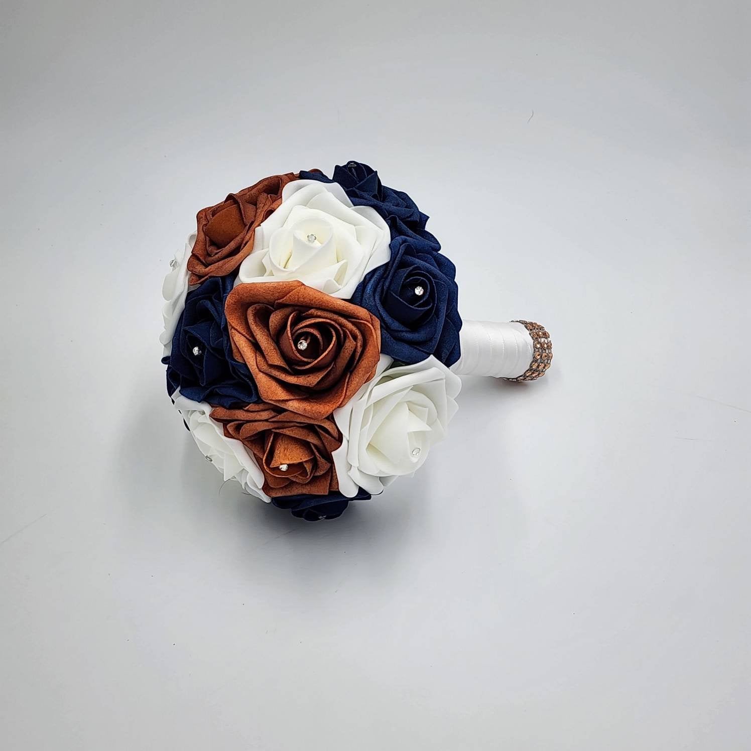 A round bridal bouquet featuring real touch roses in white, navy blue, and terracotta colors. Each rose has a small rhinestone in the center. The handle is wrapped in white satin ribbon and has a decorative jeweled band.