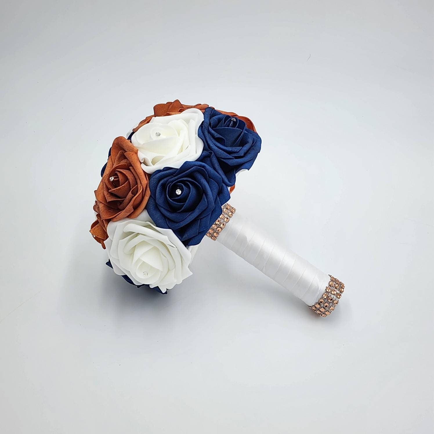 A bridal bouquet featuring artificial roses in white, navy blue, and rust orange. Each rose has a rhinestone center. The bouquet's handle is wrapped in white satin and accented with a band of rhinestones.
