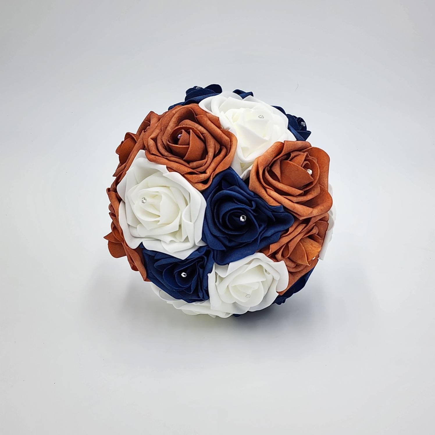 A round bouquet of artificial roses in shades of white, navy blue, and burnt orange. Each rose is adorned with a small, sparkling gem. The arrangement is tightly packed, creating a full, spherical shape, perfect for weddings.