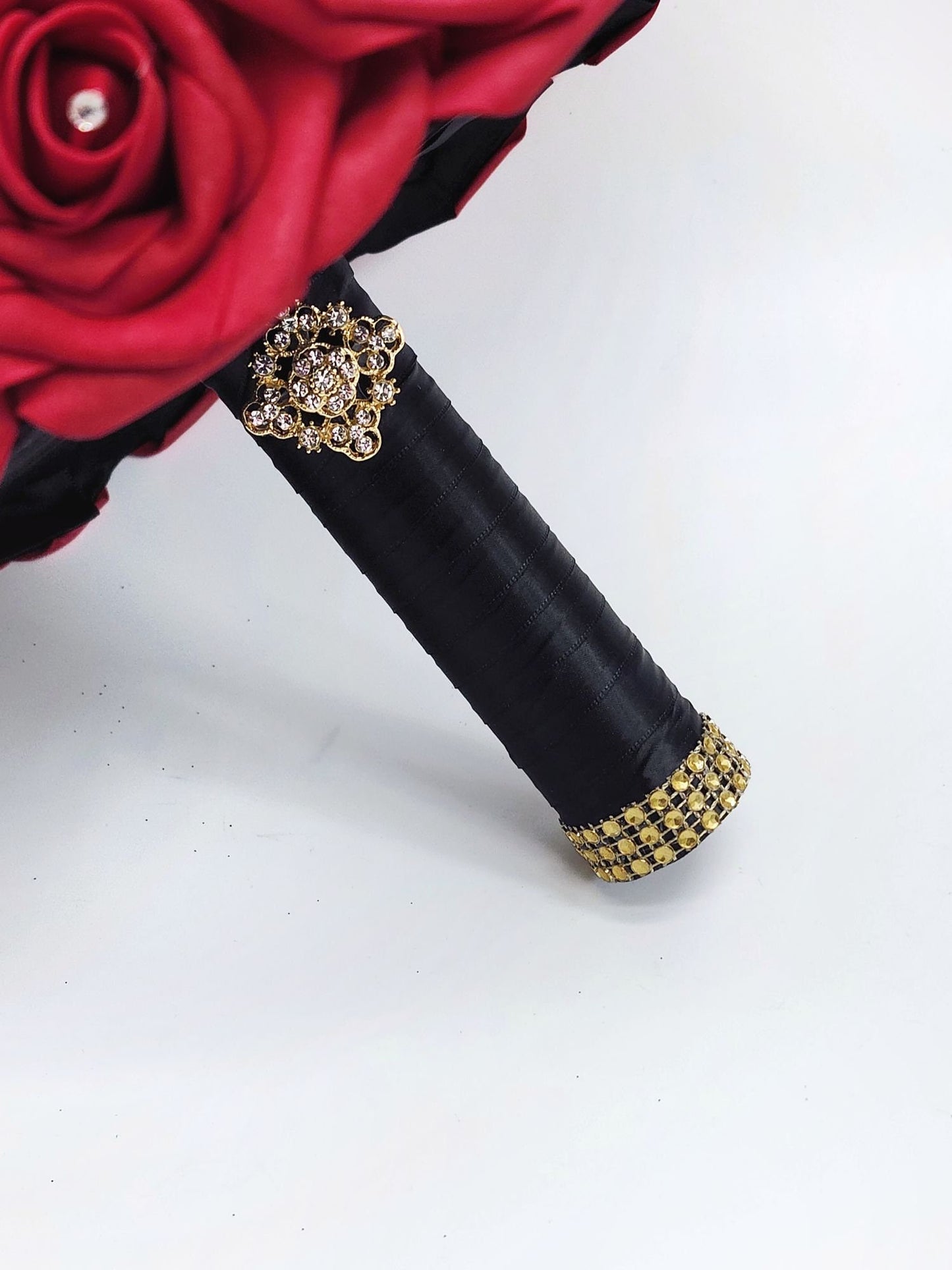 Close up of handle. Red real touch rose bouquet with black satin ribbon handle. Gold bling wrap and brooch on handle. Rhinestones on every rose.