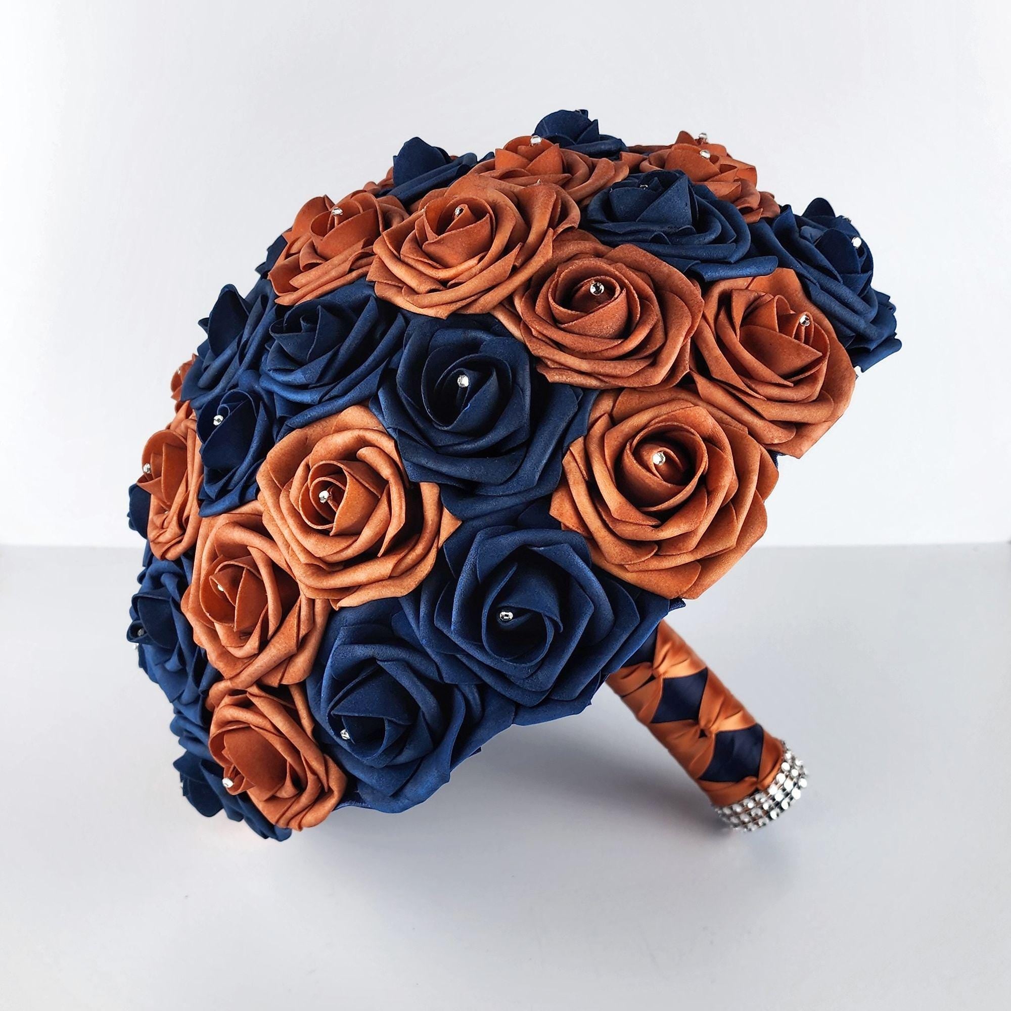 A bridal bouquet made of faux roses in navy blue and burnt orange colors. The bouquet is tied with a navy-blue ribbon and has a rhinestone embellishment.