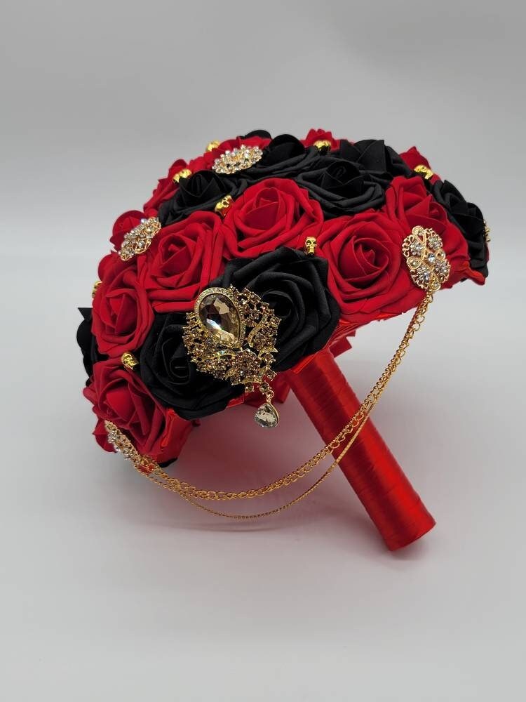 Red and black roses with gold skulls, brooches, and cascading chains. handle is made up of red satin ribbon