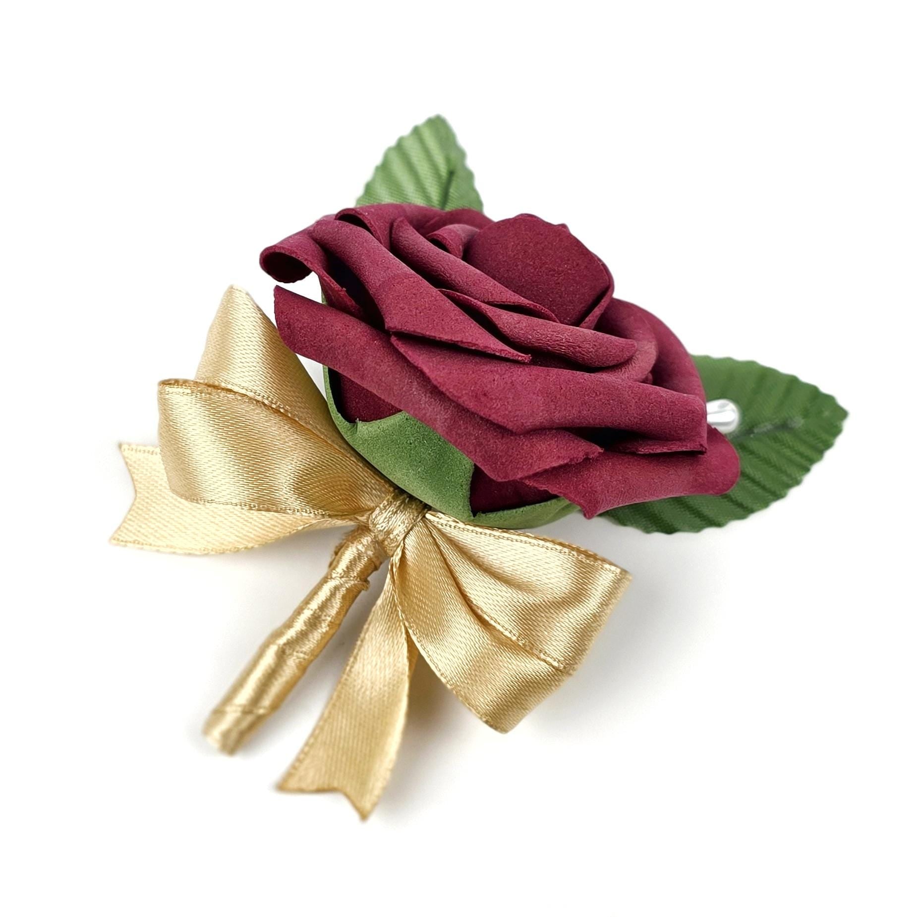 A burgundy rose boutonniere with a gold satin bow and green leaves. The flower is made of a soft material and has a small, decorative gem. This floral accessory is suitable for weddings and formal events.