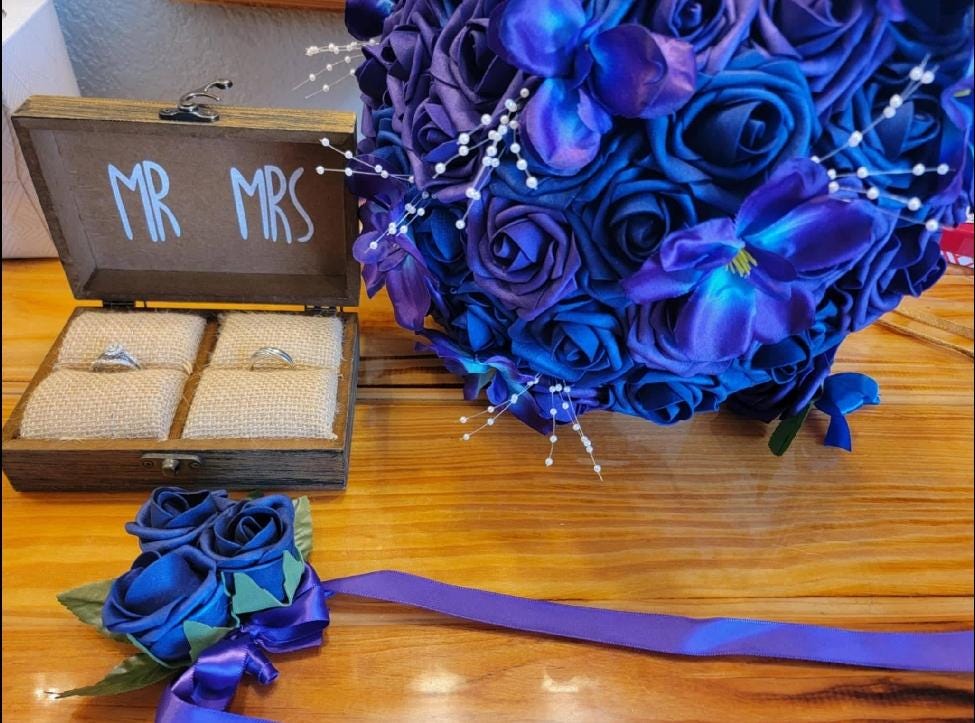 A wooden ring box with the text MR MRS, containing two rings on beige burlap cushions, next to a large bouquet of deep blue and purple artificial roses with white bead accents and a matching blue rose corsage with a purple ribbon.