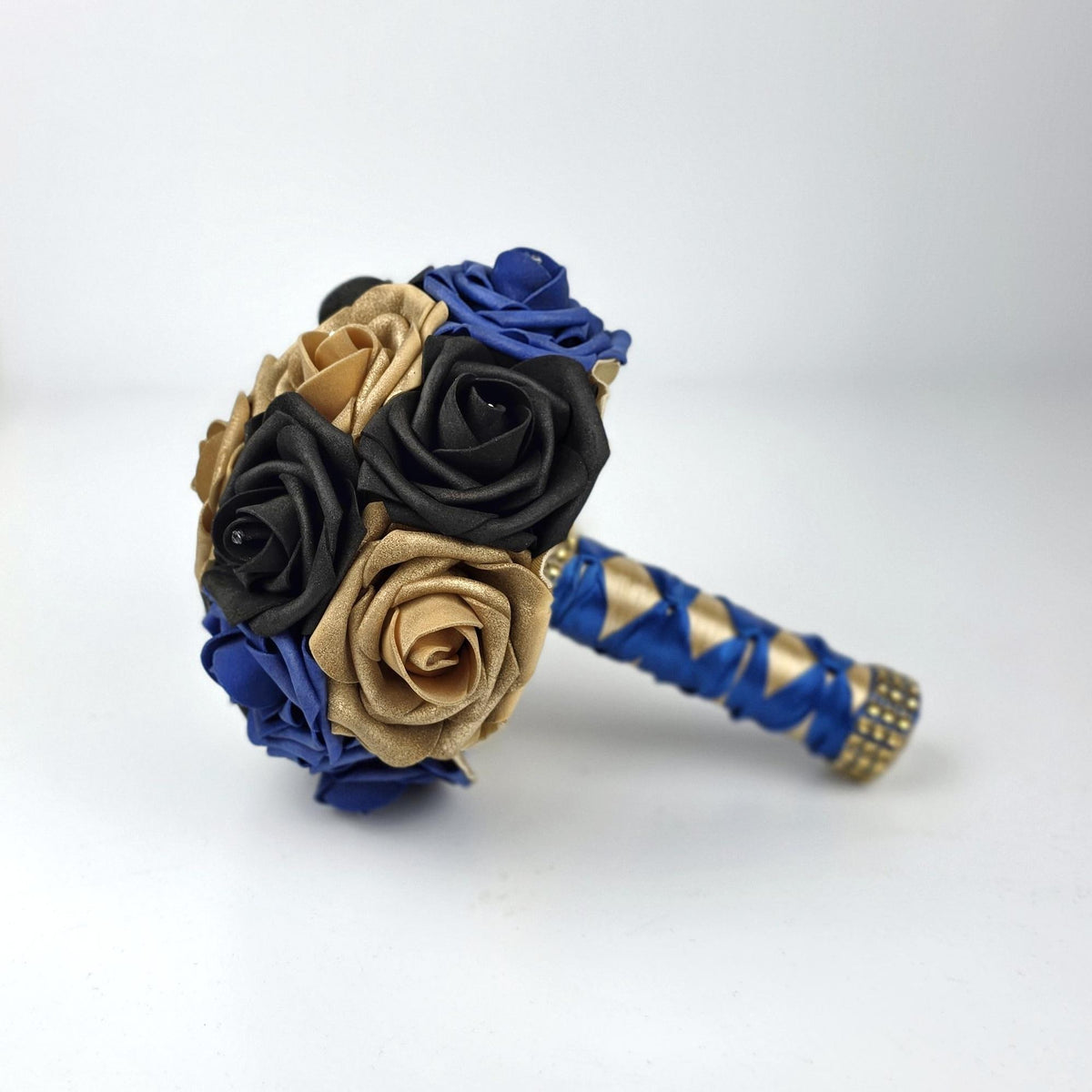A decorative bouquet featuring artificial roses in shades of royal blue, black, and metallic gold. The handle is wrapped in royal blue ribbon with a gold pattern and finished with a band of small gold-toned embellishments.