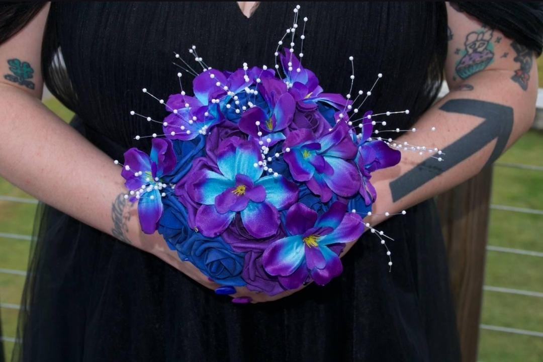 A round bouquet of artificial flowers featuring vibrant purple and bright blue petals with yellow centers. The arrangement is accented with thin, white, wire-stemmed beads that extend outward from the center of the floral cluster.
