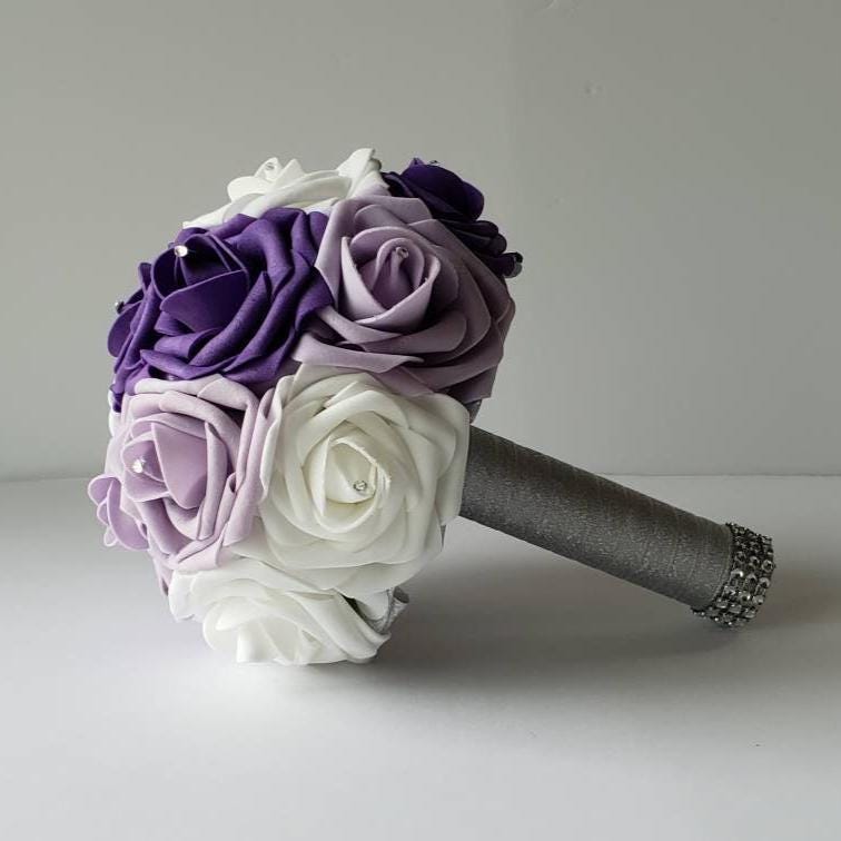 A purple, lavender, and white artificial flower bouquet with a silver ribbon and rhinestones.