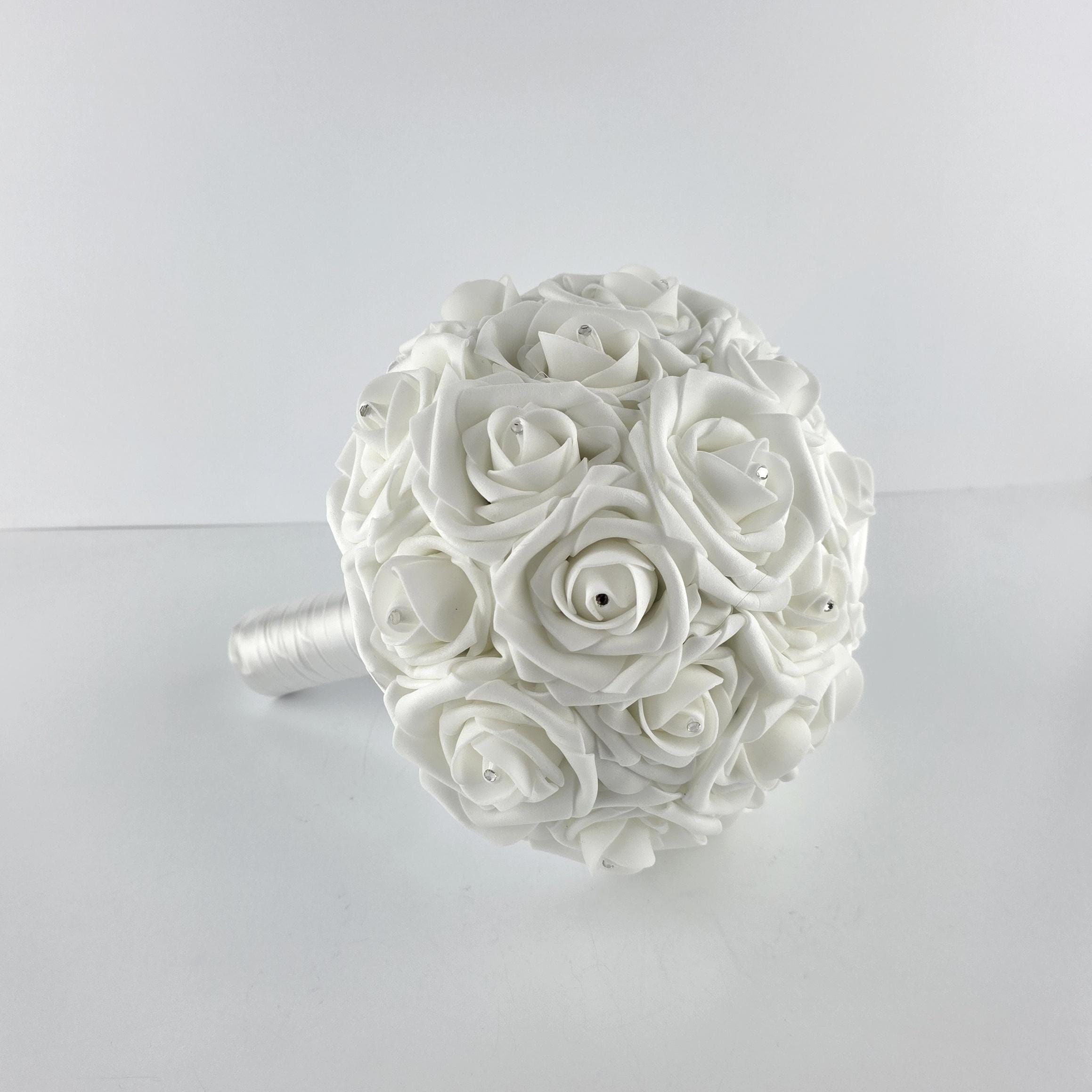 A round bridal bouquet composed of white roses with rhinestone accents. The bouquet has a white satin-wrapped handle. The roses are tightly clustered, creating a full, elegant appearance. Ideal for weddings and special events.