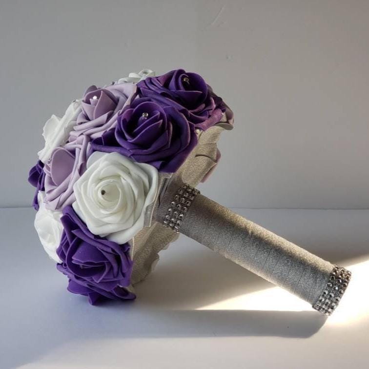 A purple, lavender, and white silk flower bridal bouquet with a silver handle and rhinestones.